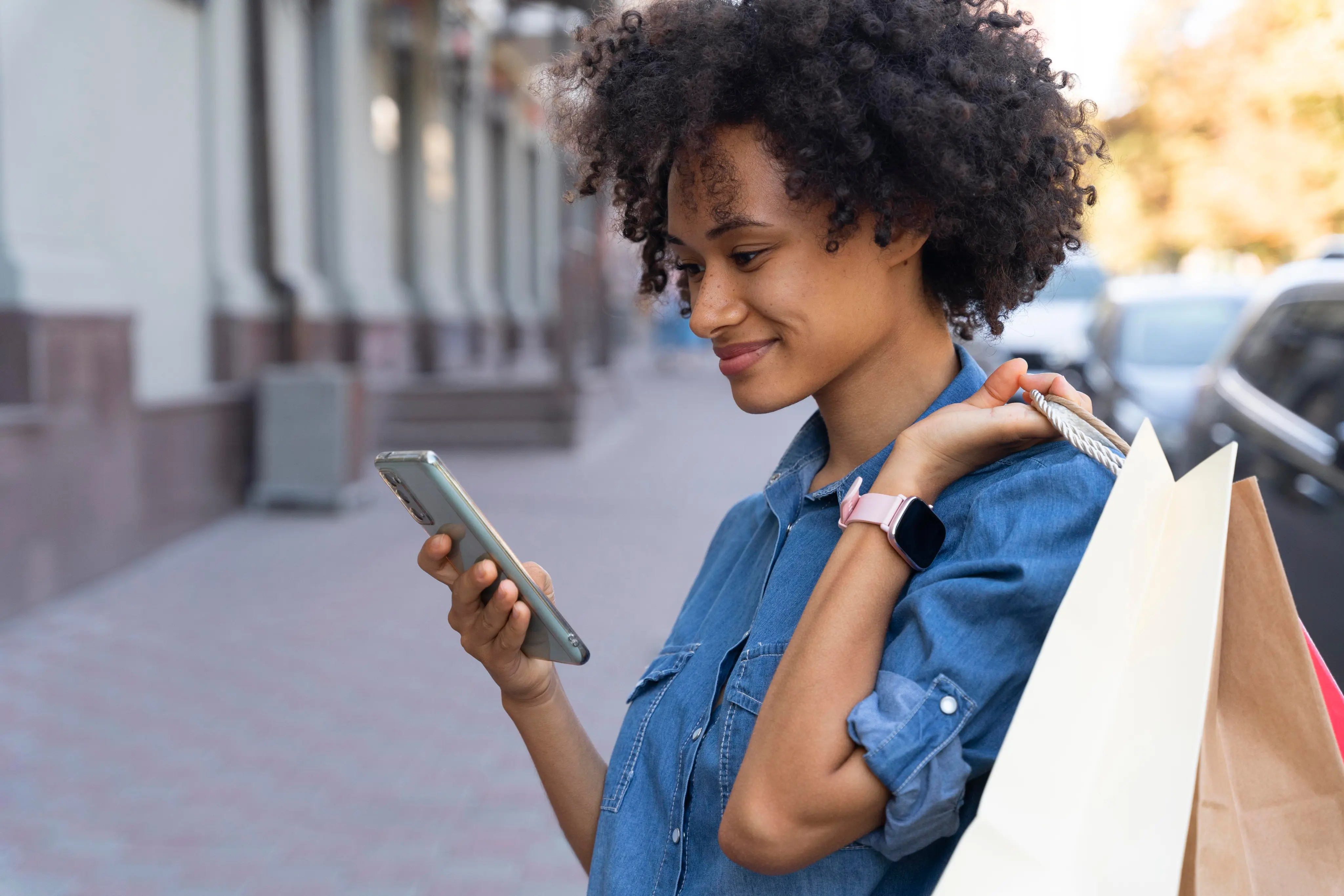a black woman smiling at her phone with a brown shopping bag over her shoulder